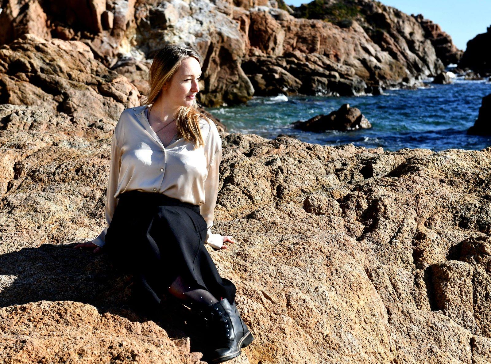 La Bàrbara Ardanuy i Queldra asseguda a les roques de la costa, amb blusa clara, mirant cap al mar blau al fons